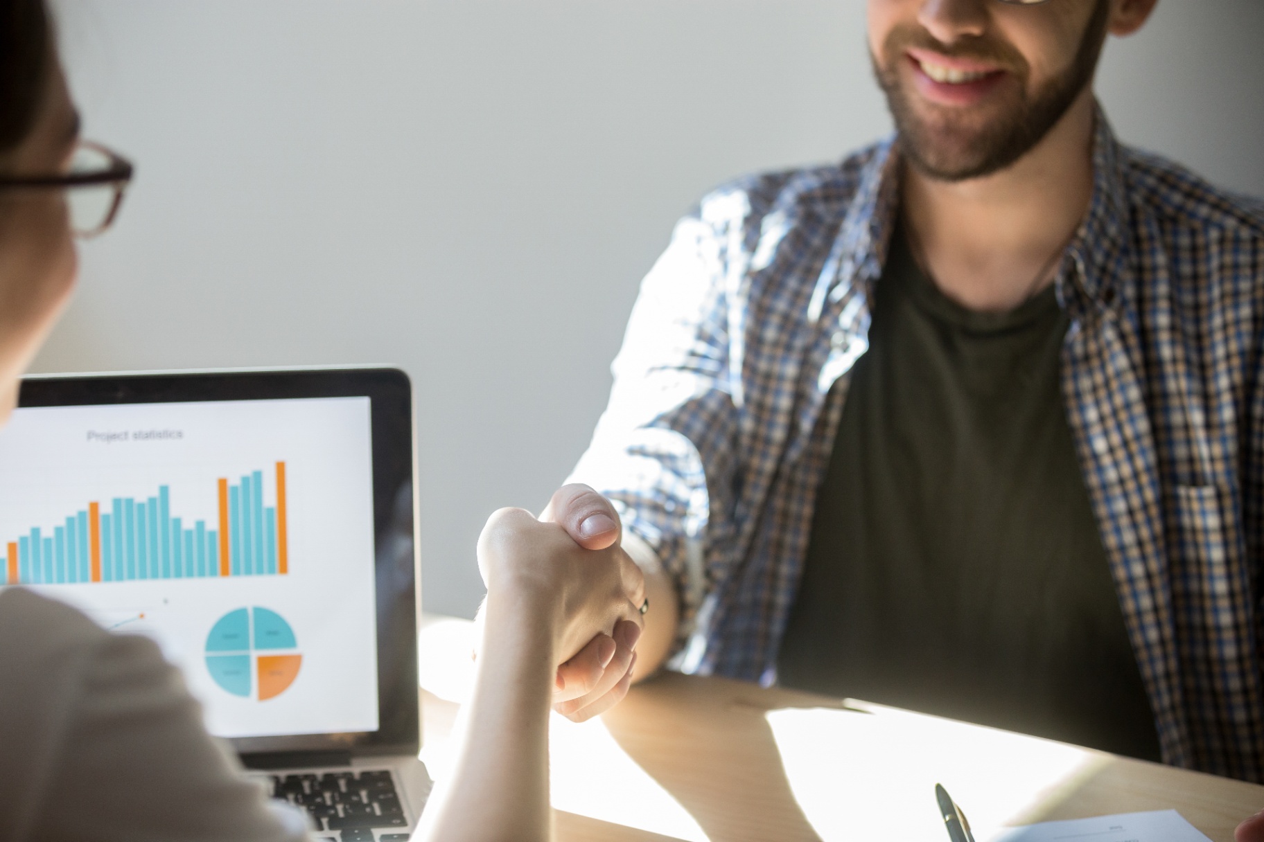 Two business people shaking hands across a laptop showing sales results.